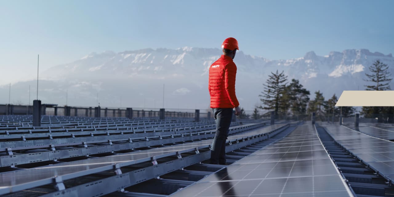 Een man met Hilti-jas en rode helm staat tussen zonnepanelen en kijkt naar een bergketen achter hem.