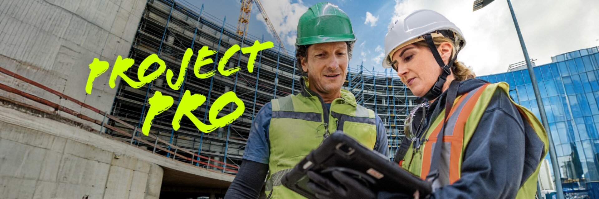 Een man en een vrouw spreken in bouwkleding over een project op de tablet, op de achtergrond is de bouwplaats zichtbaar.